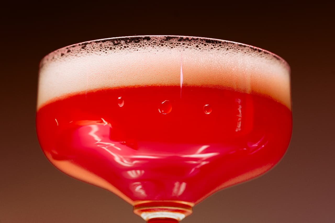 A red cocktail in a coupe glass with a pale foam cap